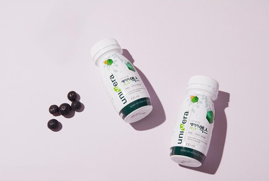 연보라 바닥에 비스듬히 놓인 에이지엑스 100 ml 병 두 개와 블랙커런트 열매