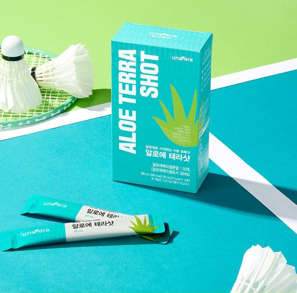 청록색 코트 위에 놓인 알로에 테라샷 상자와 액상스틱 두 포, 배드민턴 라켓과 셔틀콕