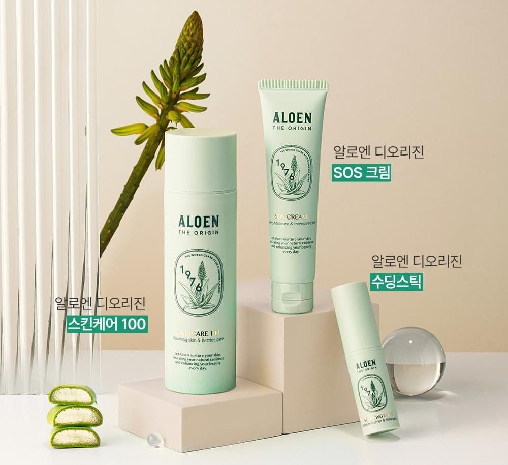 베이지색 벽과 반투명 기둥 앞, 단 위에 놓인 알로엔 디오리진 3종과 알로에 꽃대, 알로에 속살 조각, 유리 구슬. 제품 옆 이름표: 알로엔 디오리진 스킨케어 100, 알로엔 디오리진 SOS 크림, 알로엔 디오리진 수딩스틱