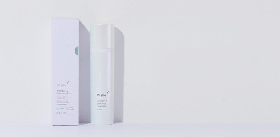 연보랏빛 회색 벽 앞 선반 위의 W389 더마 브라이트닝 토너 상자(흰 · 연보라)와 토너 병
