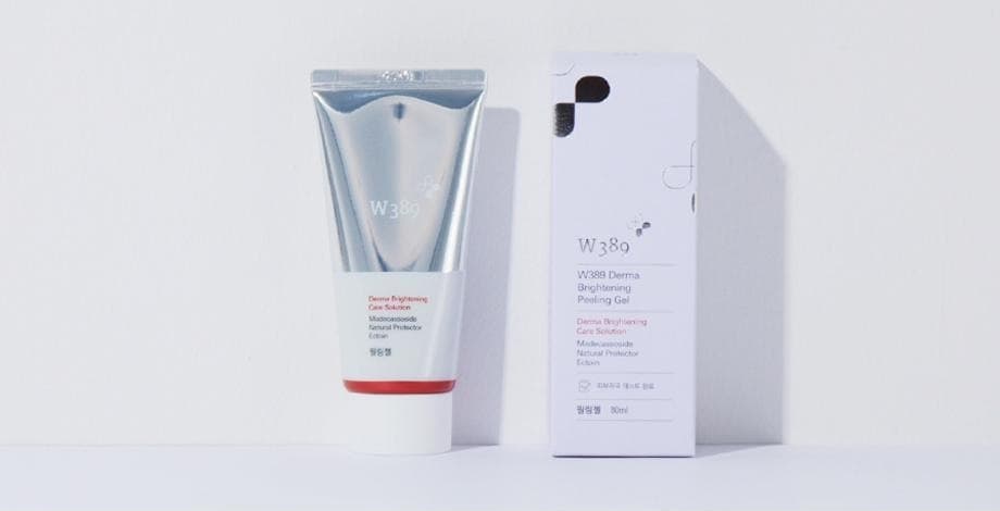 흰 벽 앞 선반 위에 선 W389 더마 브라이트닝 필링젤 튜브와 상자. 상자 글자: W389, W389 Derma Brightening Peeling Gel, Derma Brightening Care Solution, Madecassoside Natural Protector Ectoin, 필링젤 80ml