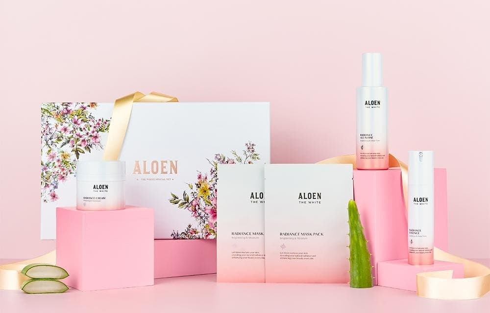 분홍 받침 위 꽃무늬 기프트 박스(ALOEN, 금빛 리본)와 알로엔 더화이트 래디언스 크림 단지 · 마스크팩 파우치 둘 · 올인원 병 · 에센스 병, 알로에 잎