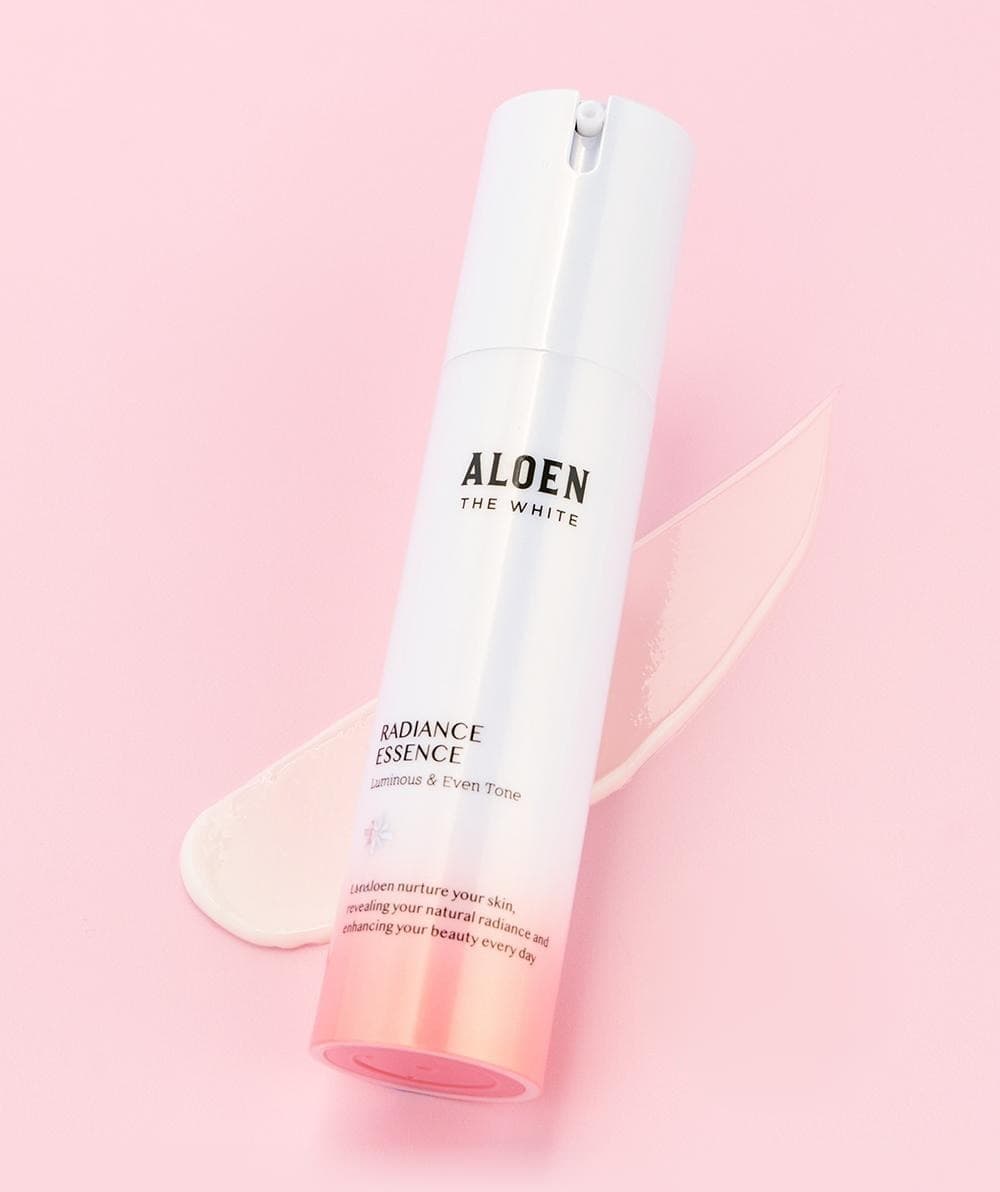 분홍 지면 위 비스듬히 선 알로엔 더화이트 래디언스 에센스 병과 그 뒤로 길게 펴 바른 크림 텍스처. 병 글자: ALOEN THE WHITE, RADIANCE ESSENCE, Luminous & Even Tone, Let Aloen nurture your skin, revealing your natural radiance and enhancing your beauty every day