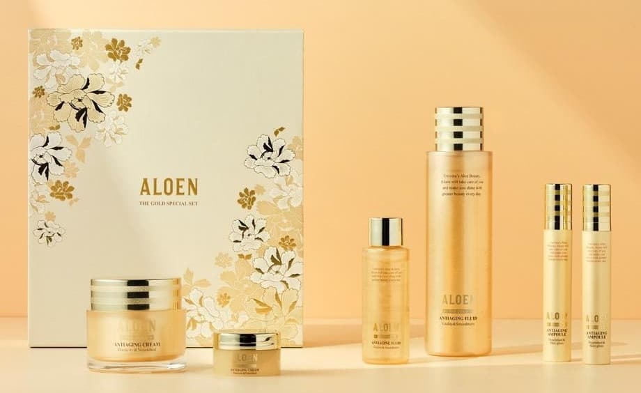 금박과 흰색 모란 무늬의 아이보리 선물 상자(ALOEN, THE GOLD SPECIAL SET) 앞에 알로엔 안티에이징 크림 50ml 와 10ml, 오른쪽에 플루이드 50ml 와 200ml, 앰플 2개가 금빛 벽과 탁자 위에 놓여 있다. 용기 글자: ALOEN, THE GOLD, ANTIAGING CREAM Elasticity & Nourished, ANTIAGING FLUID Vitality & Smoothness, ANTIAGING AMPOULE Nourished & Skin gloss