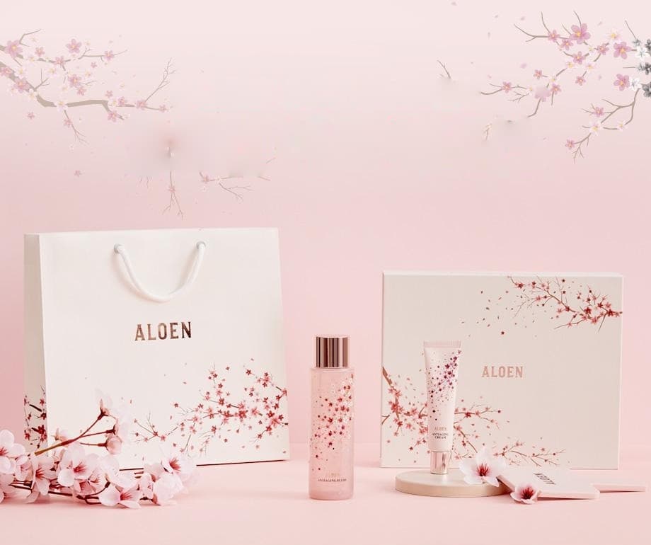 분홍 벽 앞 벚꽃 가지 아래, 벚꽃 무늬 흰 쇼핑백(ALOEN) · 벚꽃 무늬 알로엔 안티에이징 플루이드 병 · 안티에이징 크림 튜브 · 벚꽃 무늬 선물 상자(ALOEN) · 둥근 받침과 손거울 · 벚꽃 송이. 용기 글자: ALOEN, ANTIAGING FLUID, ANTIAGING CREAM