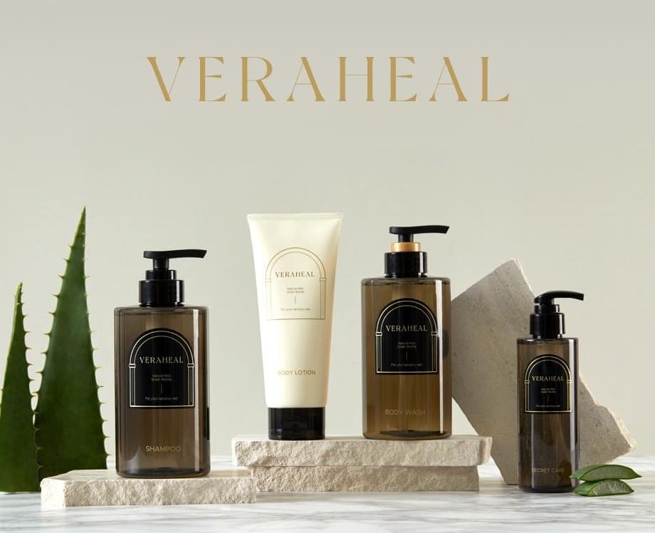 회베이지 벽 위쪽의 금색 VERAHEAL 워드마크. 아래는 알로에 잎과 돌 받침 위 베라힐 라인 넷 — SHAMPOO · BODY LOTION · BODY WASH · SECRET CARE