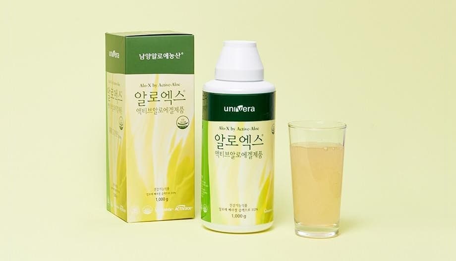 연두 벽 앞의 알로엑스 상자(남양알로에농산 · Alo-X by Active-Aloe · 알로엑스 · 액티브알로에겔제품 · 1,000 g)와 흰 뚜껑 병(univera · 알로엑스), 알로에 음료가 담긴 잔