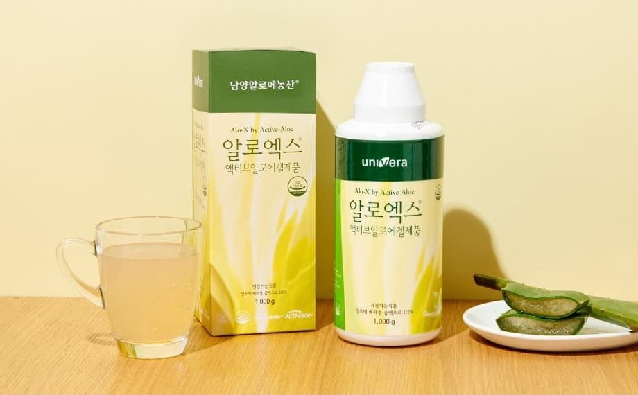 나무 탁자 위의 알로엑스 상자와 병, 알로에 음료가 담긴 유리컵, 접시 위의 알로에 잎